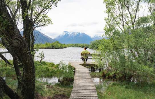 Man sits on a seat overlooking a lake, surrounded by green trees with a snowcapped mountain in the background