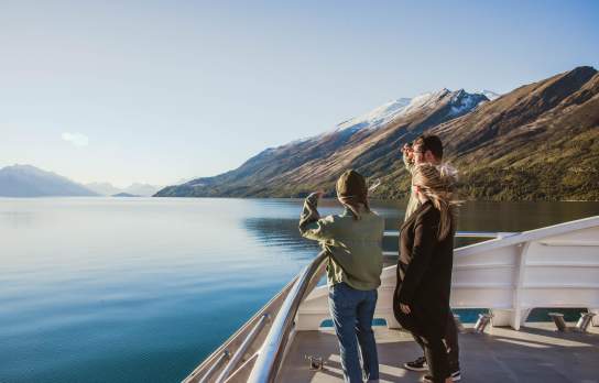 Three people standing on the deck of a boat, gazing out at the calm blue waters of a lake surrounded by mountains. The landscape features snow-capped peaks and lush greenery under a clear blue sky, creating a serene and picturesque setting