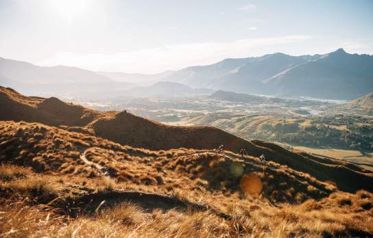 Far shot of three bikers descending a golden tussock-covered mountain with valley and mountain in the background