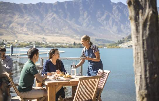 Couple being served by a waitress outside at the Boat Shed café, with the Remarkables Mountains in the background