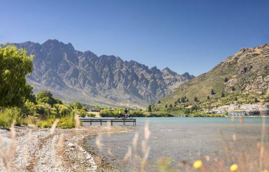 Two people having a rest on a pier looking out over the lake and Remarkables mountains while biking the Frankton Trail