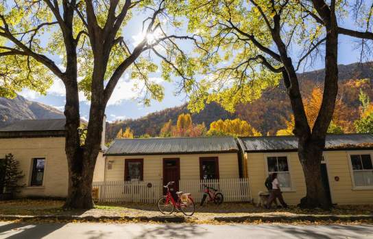 Historic miners’ cottages in Arrowtown dappled in autumn sunlight, framed by golden trees, with red bikes parked out front and a couple walking a dog beneath the canopy.