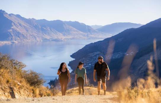 Three people walking on a steep track with mountains and lake views in the background