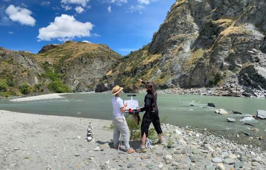 Two people standing beside a braided alpine river surrounded by rugged mountains and rocky cliffs under a bright blue sky.
