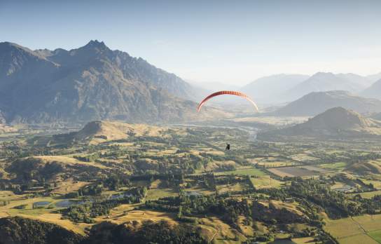 Person paragliding over a green valley with the Remarkables mountain range in the background