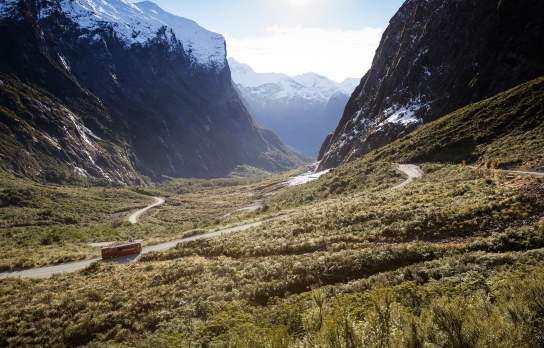 Wide shot of a bus traveling on the road to Milford Sound surrounded by towering snow-capped mountains and native bush