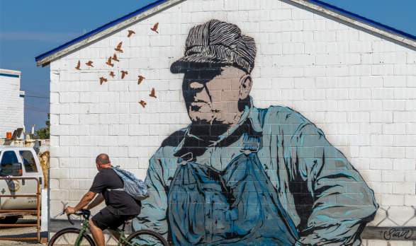 A man biking past a mural of an older working man titled Keep em Rollin by Chris Stain from the BOOM! Salina festival in Salina, Kansas