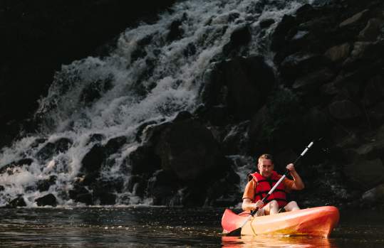 a man wearing a life-jacket and kayaking with a waterfall in the background