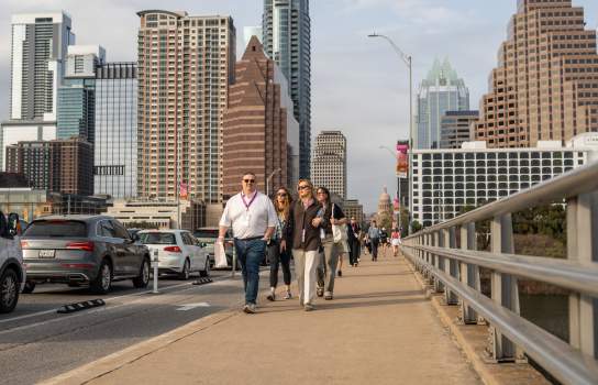 A group of people wearing SXSW badges walk across the Congress Avenue Bridge with the Austin skyline in the background.