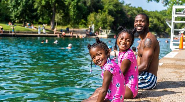 Two little girls and their father sit on the edge of the pool and smile at Barton Springs