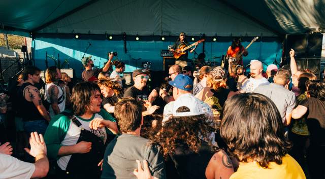 Crowd of people watching a band on at outdoor stage on a sunny day.