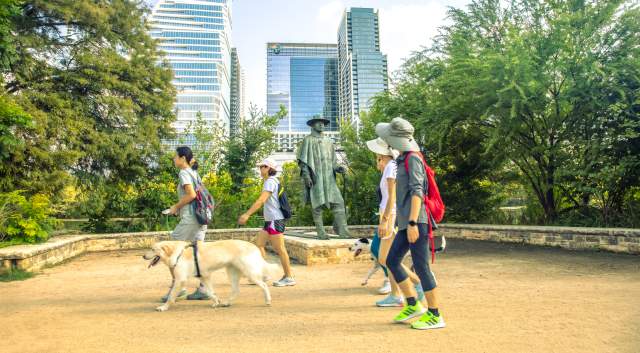 Men and women walking past the Stevie Ray Vaughan statue on the Butler Hike and Bike Trail with their dogs.