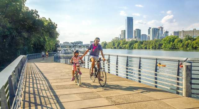 Image of a man and daughter riding bikes on the Butler Hike and Bike trail boardwalk with Austin Downtown in the background.