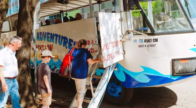 A woman, man and child loading on to the Austin Duck Adventure tour bus, outside of the Austin Visitor Center.
