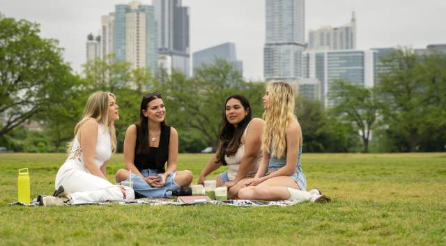 Image of four women sitting on a blanket in Zilker Park chatting with the Austin skyline in the background.