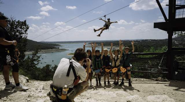 Image of a man taking a group photo of kids at Lake Travis Zipline with someone zipping down behind them.