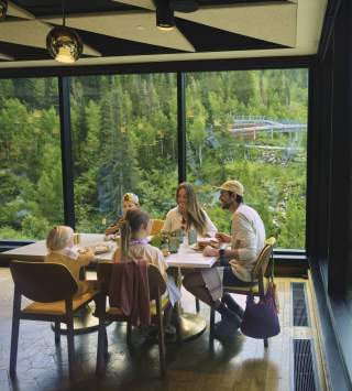 Family dining at an indoor table with very large glass windows all around looking out at the side of a mountain covered in green pine trees