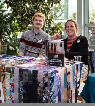 Vendor table covered in a colorful cloth that says UMFA, has swag on it and 2 smiling people sitting behind it.