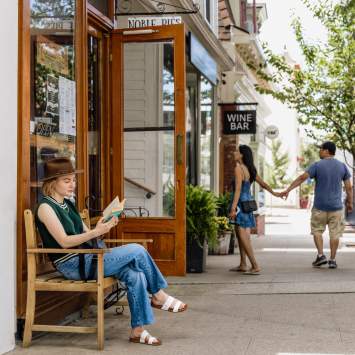 A woman sits on a bench reading a book while a couple in the background is walking into a shop.