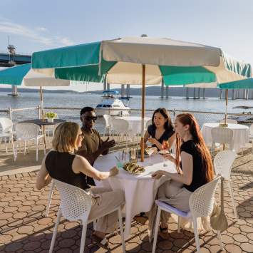 A group of four people sit at an outdoor dining table overlooking the Hudson River. They are laughing and chatting.