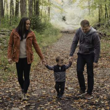A family of three holds hands and walks along a leaf-strewn trail.