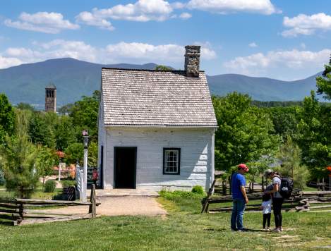 Historic white cabin in Page County, Virginia with lush greenery, mountain backdrop, and visitors exploring
