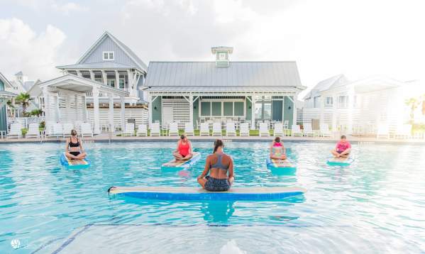 A woman with crossed legs sits atop a surf board floating in a pool, back to the camera. Four women, also with crossed legs, sit atop surf boards facing the instructor.