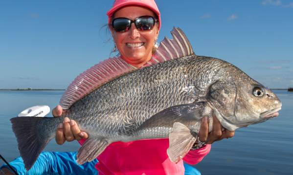 A woman in a pink shirt and hat smiling while holding a large black drum fish on a boat.