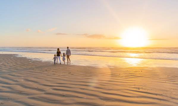 A family of four walks on the beach away from the camera towards the left. The sun is rising to the right.