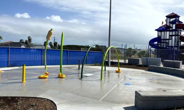 On a gray pad sits several green and yellow hoops and poles with water for kids to splash on. A playground is in the back right.
