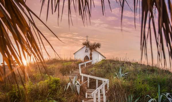 On top of a grassy hill at sunset sits a small white chapel topped with a cross. Steps with a white railing lead up to the chapel and palm tree fronds frame the photo