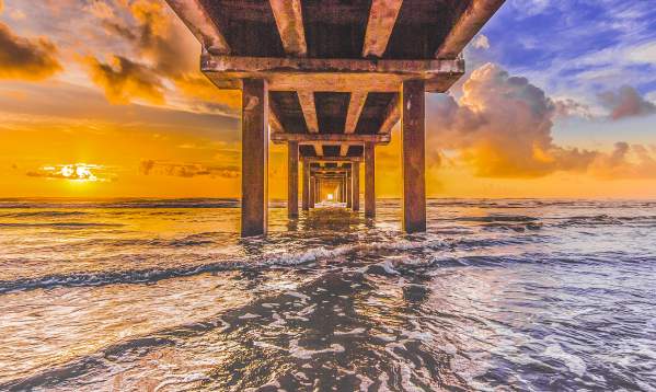 The sun rises just over the water from the perspective of under a pier