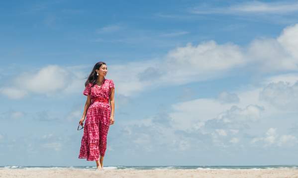 A woman in a pink and red dress walks along the beach