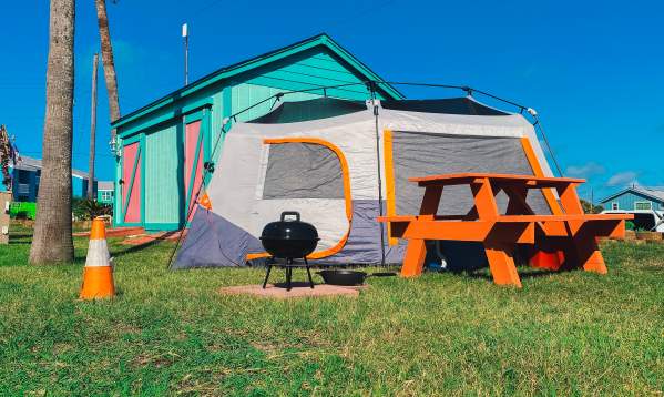 Gray and orange tent sits on grass next to an orange picnic table and black camp grill