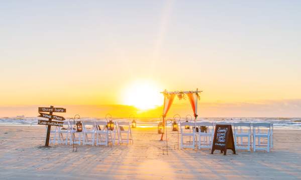 The sun rises on a wedding ceremony setup on the beach. There are rows of white chairs, wooden signs, and a tulle/floral arch.