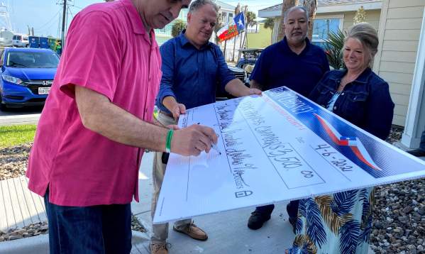 A man in a pink shirt signs a giant check while three others hold it up.