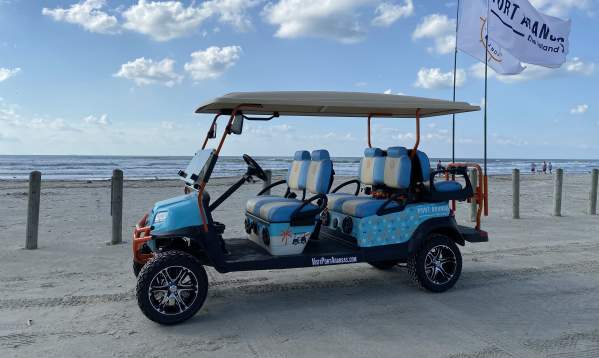 A light blue and patterned golf cart with two large flags sits on the beach.