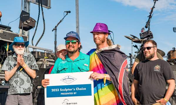 Two men, one draped in multicolored flags and a purple fedora and the other in a teal fishing shirt and hat, pose with a sign that reads "2023 Sculptor's Choice"