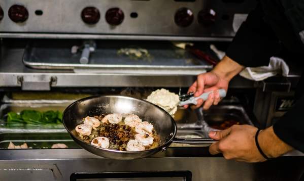 A pair of hands holds out a pan full of sizzling shrimp over an oven while scooping another ingredient into the pan