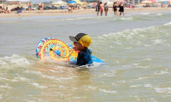 A child in a ball cap swims with a tie-dye boogie board near the beach