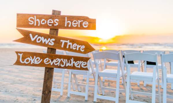 A brown sign is stuck in the sand. It reads "Shoes here," "Vows there," and "Love everywhere."
