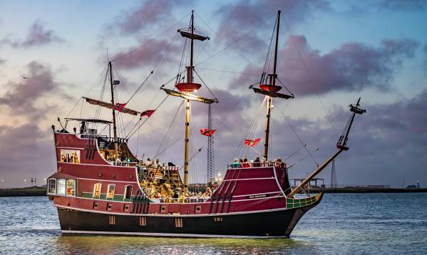 Large red pirate ship in the ocean with lights on in facing the deck. The sun has almost set and gives the sky and clouds a purple tint.
