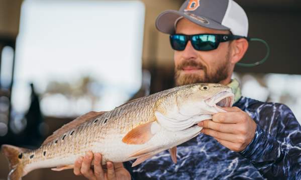 A smiling man in a cap and sunglasses holds up a speckled trout