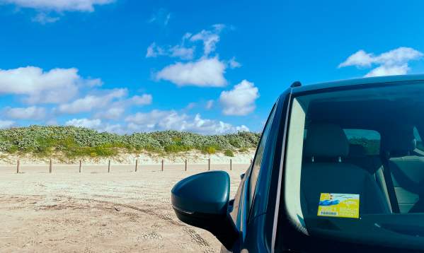 The left side shows blue sky with dunes and sand, right side is the front of a car with a yellow sticker on the windshield