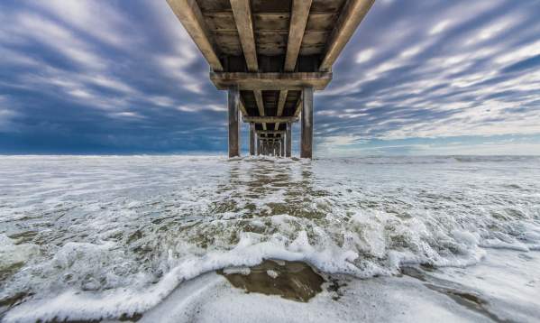 Foamy seawater rushes underneath a pier with a bright, cloudy sky