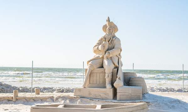 Sand sculpture of a pirate drinking out of a cup and sitting on barrels. In front of him is a rowboat made of sand