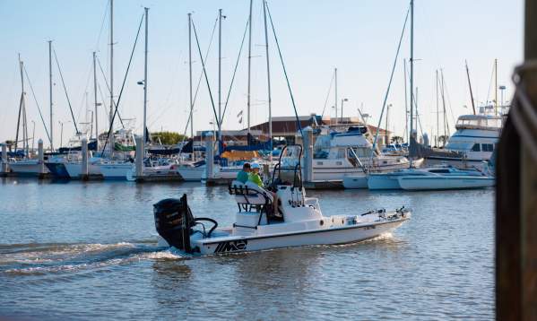A fishing boat with two people in the driver's seat putters through a marina with rows of sailboats behind