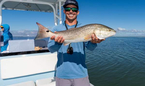 A man in a blue long-sleeve shirt and sunglasses smiling while holding a large redfish on a boat.