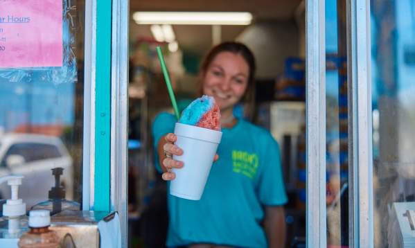 Girl in turquoise shirt holds out a red and blue snow cone in a styrofoam cup outside a window