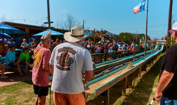Two men stand in front of narrow wooden racing tracks with belt sanders while a crowd watches